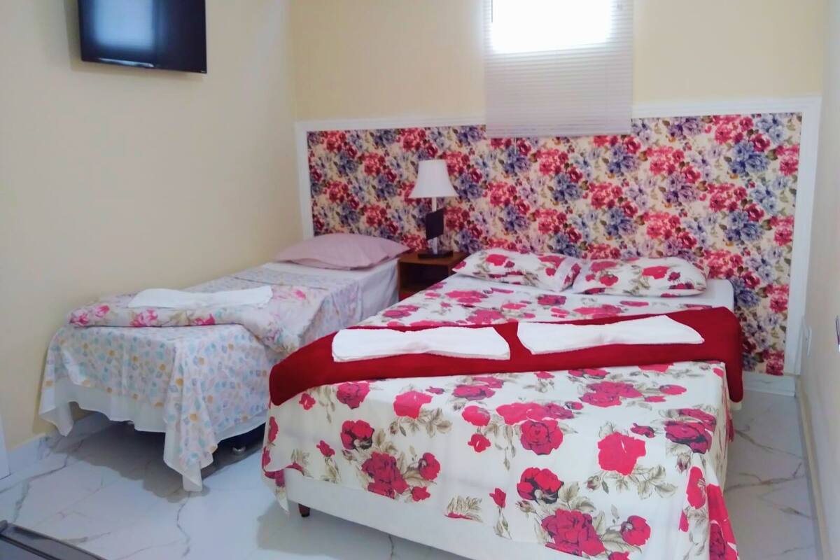 A bedroom with two beds, one larger with a floral headboard and one smaller with a plain headboard. There is a TV mounted on the wall and a window with blinds.