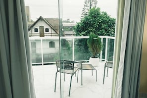 Un balcón con una planta en maceta y dos sillas, con vista a una zona residencial con casas y un árbol grande.