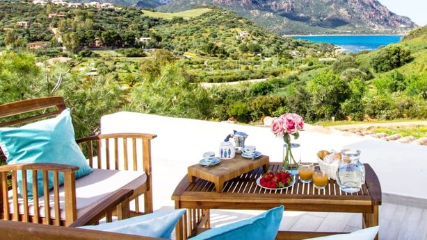 A wooden table set with breakfast items, overlooking a coastal landscape with greenery and a view of the sea.