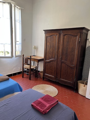 A room with two beds, a wooden wardrobe, a table with a lamp, and a view through a window.