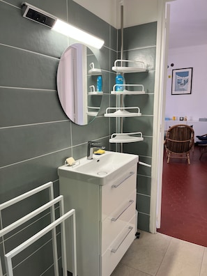 Un baño con un lavamanos blanco, espejo y repisas con artículos de aseo.