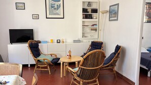 A living room with a flat-screen TV, wicker chairs, a white bookshelf, and a dining area with a table. - Detached villa in residential area, beach and shops nearby (Sanary-sur-Mer)