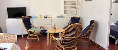 A living room with a flat-screen TV, wicker chairs, a white bookshelf, and a dining area with a table.