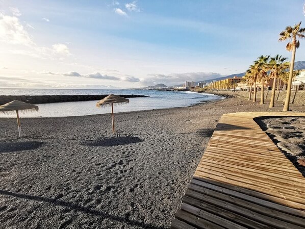 A beach with a wooden walkway, palm trees, and a rocky shoreline. - Home2Book Caletillas Breeze & Comfort (Tenerife)