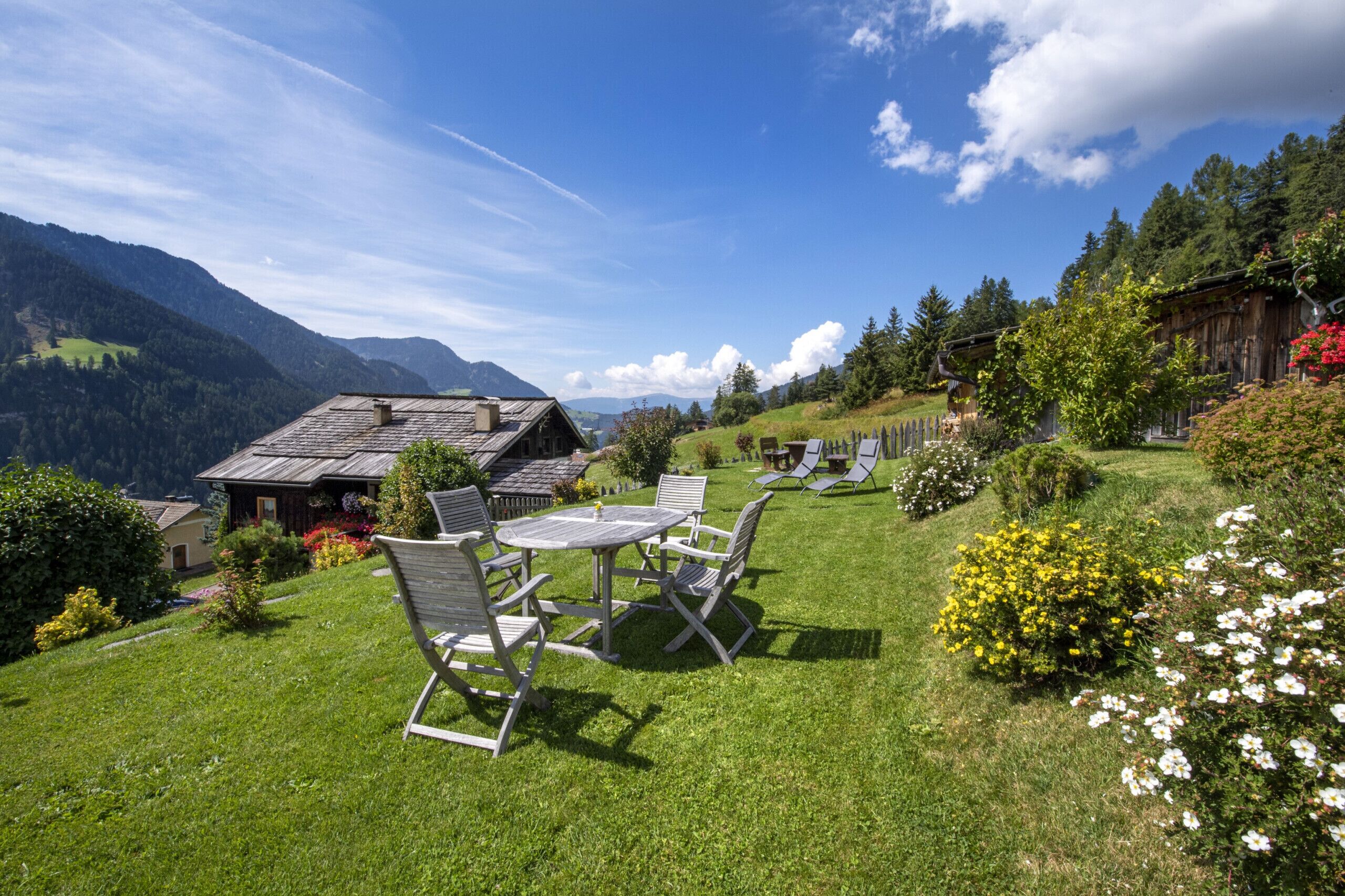 Un'area salotto esterna rustica con tavolo e sedie, immersa nel verde e circondata da fiori, con montagne e un cielo sereno sullo sfondo.