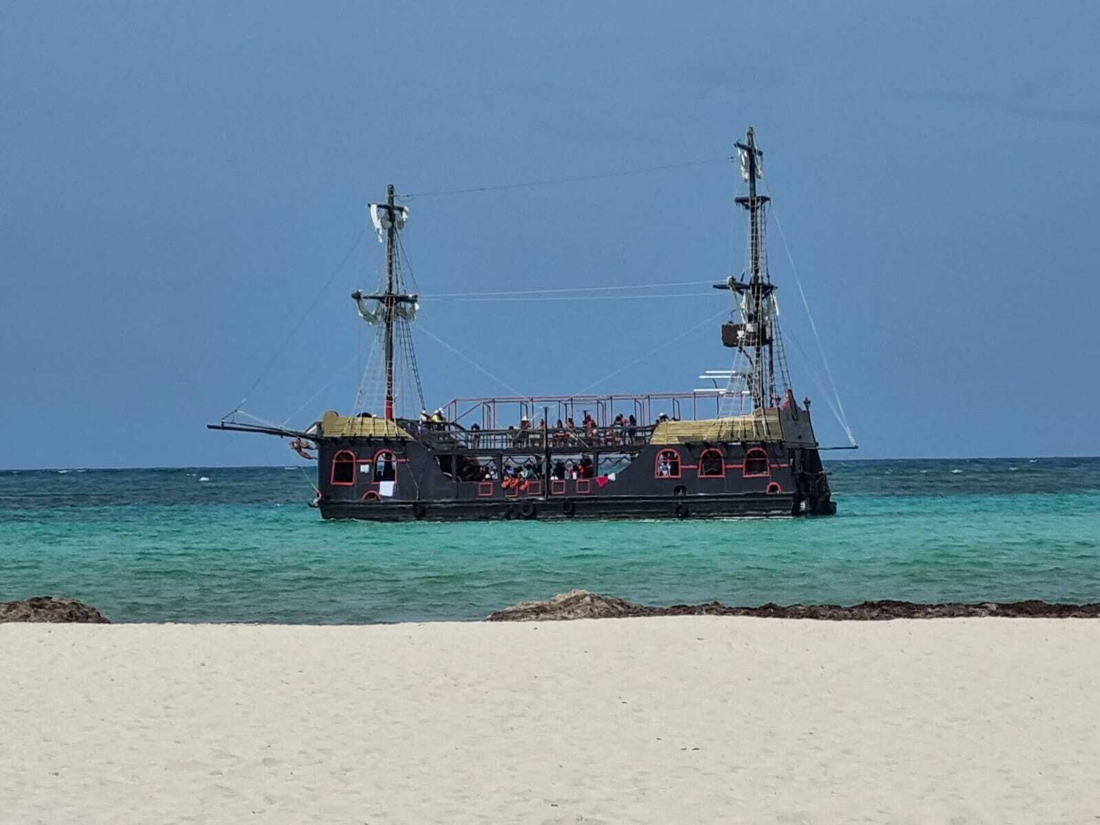 Een groot, zwart schip met meerdere dekken en masten, afgemeerd aan een zandstrand.