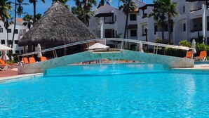 Una piscina con estructura de techo de paja, rodeada de sillas de descanso anaranjadas y palmeras.