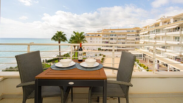 A balcony with a table set for two, overlooking a beach and a hotel building.