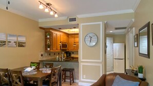A dining area with a table and chairs, a kitchen with wooden cabinets, a clock, and a refrigerator. - Gorgeous Ground Floor Condo in World Class Resort, Pool, Spa, Amenities Galore! (Naples)