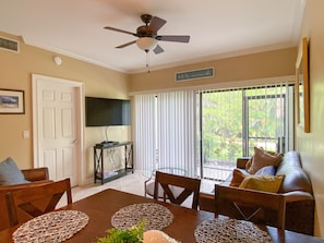 A living room with a ceiling fan, a television, a sofa, and a dining table with placemats. - Gorgeous Ground Floor Condo in World Class Resort, Pool, Spa, Amenities Galore! (Naples)