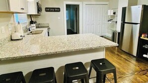 A kitchen with granite countertops, black stools, a stainless steel refrigerator, and a sign on the wall. - Greer Twin Cottage A - Walk to City Park and Downtown Greer (Greer)