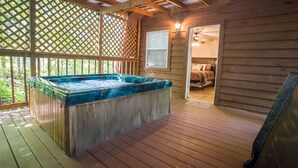 A wooden deck with a hot tub, a covered patio area, and a bedroom visible through an open door. - Rock Right Inn* Cozy /Mtn Views*Creek*Firepit*Hot tub*1.5 acres* 4bed/2 bed*8-10 (Cosby)