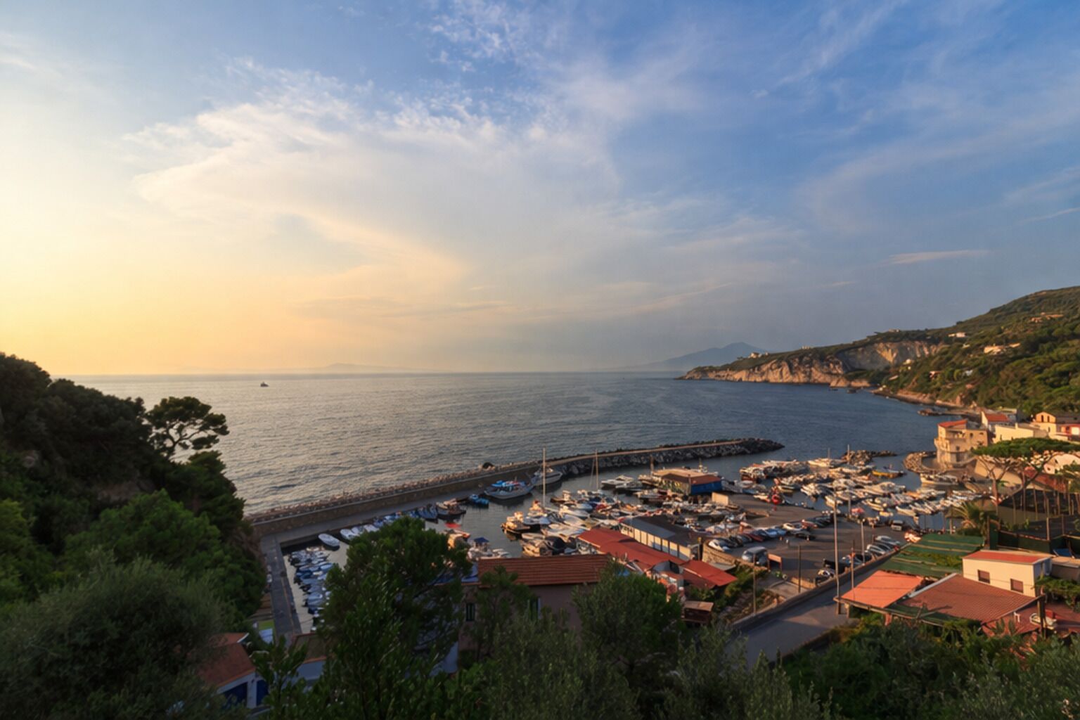 A coastal marina with boats docked, a pier extending into the water, and buildings with red roofs.