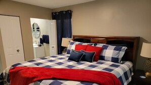 Una cama bien hecha con colcha a cuadros y manta roja, un cabecero de madera, una mesita de noche con lámpara y un espejo en la pared.