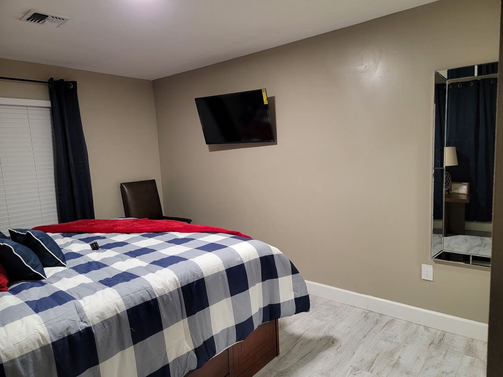 Un dormitorio con una cama, un televisor montado en la pared, una silla y una ventana con cortinas.