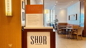 Un hall d’hôtel comprenant un comptoir d’accueil portant l’inscription « SHOR », un espace repas avec des tables et des chaises, et un panneau mural affichant des informations sur le bien-être des clients.