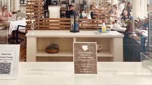 Un restaurant avec des lambris en bois, une enseigne indiquant « Beachhouse at the Moana » et proposant des steaks, des grillades de fruits de mer et des boissons alcoolisées, ainsi qu’une autre enseigne indiquant « The Veranda » et offrant des petits-déjeuners, des déjeuners, des collations de l’après-midi, et un code QR pour le traçage des contacts.
