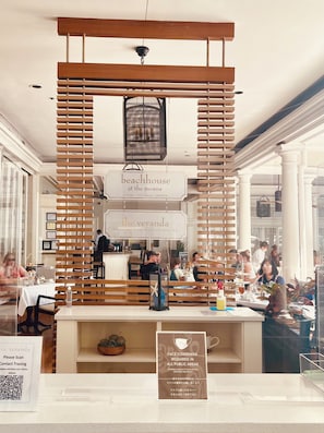 Ein Restaurant mit Holzhakobarbeiten, ein Schild für 'beachhouse at the moana' mit Angeboten wie Steaks, Meeresfrüchte-Grill und Spirituosen, und ein weiteres Schild für 'the veranda' mit Frühstück, Mittagessen, Nachmittagstee und einem QR-Code zur Kontaktverfolgung.