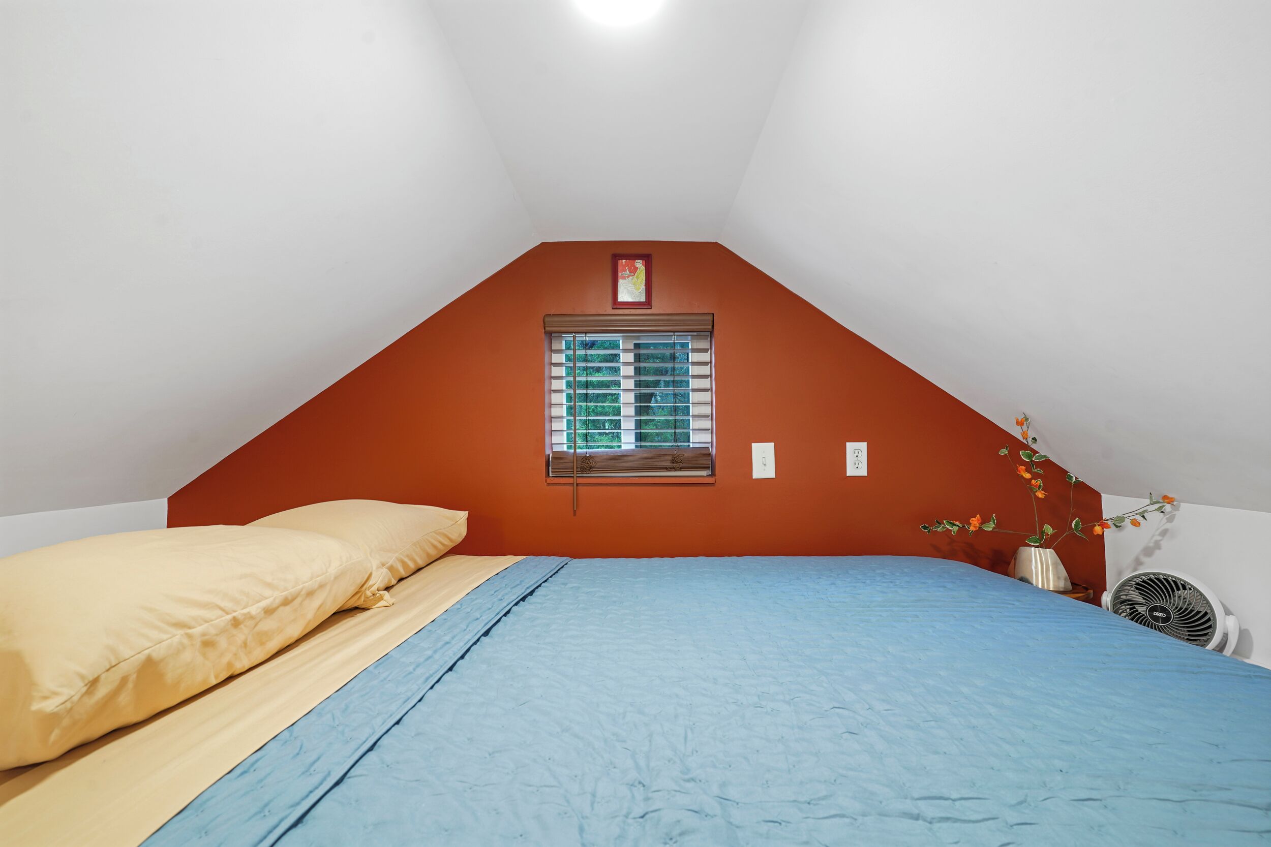 Quarto com teto inclinado, janela com persianas, cama com roupa de cama azul e bege, mesinha com um ventilador e um vaso com flores.