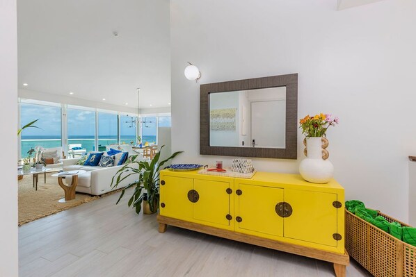 Uma sala de estar luminosa com um armário amarelo, um espelho grande e vista para o oceano.