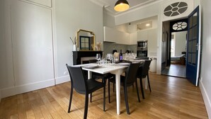 Un coin repas avec une table blanche dressée pour quatre personnes, des chaises noires, un luminaire suspendu noir et une cuisine en arrière-plan.
