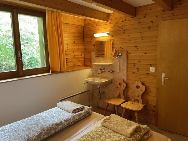 Una habitación pequeña y acogedora con paredes de madera, una cama individual, una mesita de noche, una silla de madera y un lavabo integrado, además de un espejo.