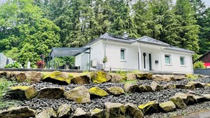 A white house with a covered patio, surrounded by a rocky garden and dense forest.