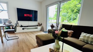 A modern living room with a flat-screen TV, a sofa, a coffee table, and a dining area with a chair and table.