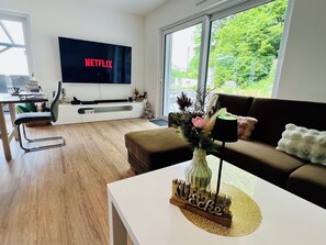 A modern living room with a flat-screen TV, a sofa, a coffee table, and a dining area with a chair and table.