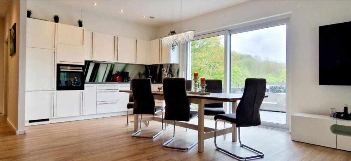Une cuisine moderne avec des meubles blancs, un coin repas avec des chaises noires et une grande fenêtre offrant une vue sur la verdure.