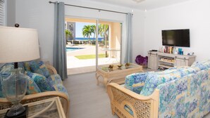 Un salon avec un canapé, une table basse, un téléviseur et une vue sur une piscine et des palmiers.