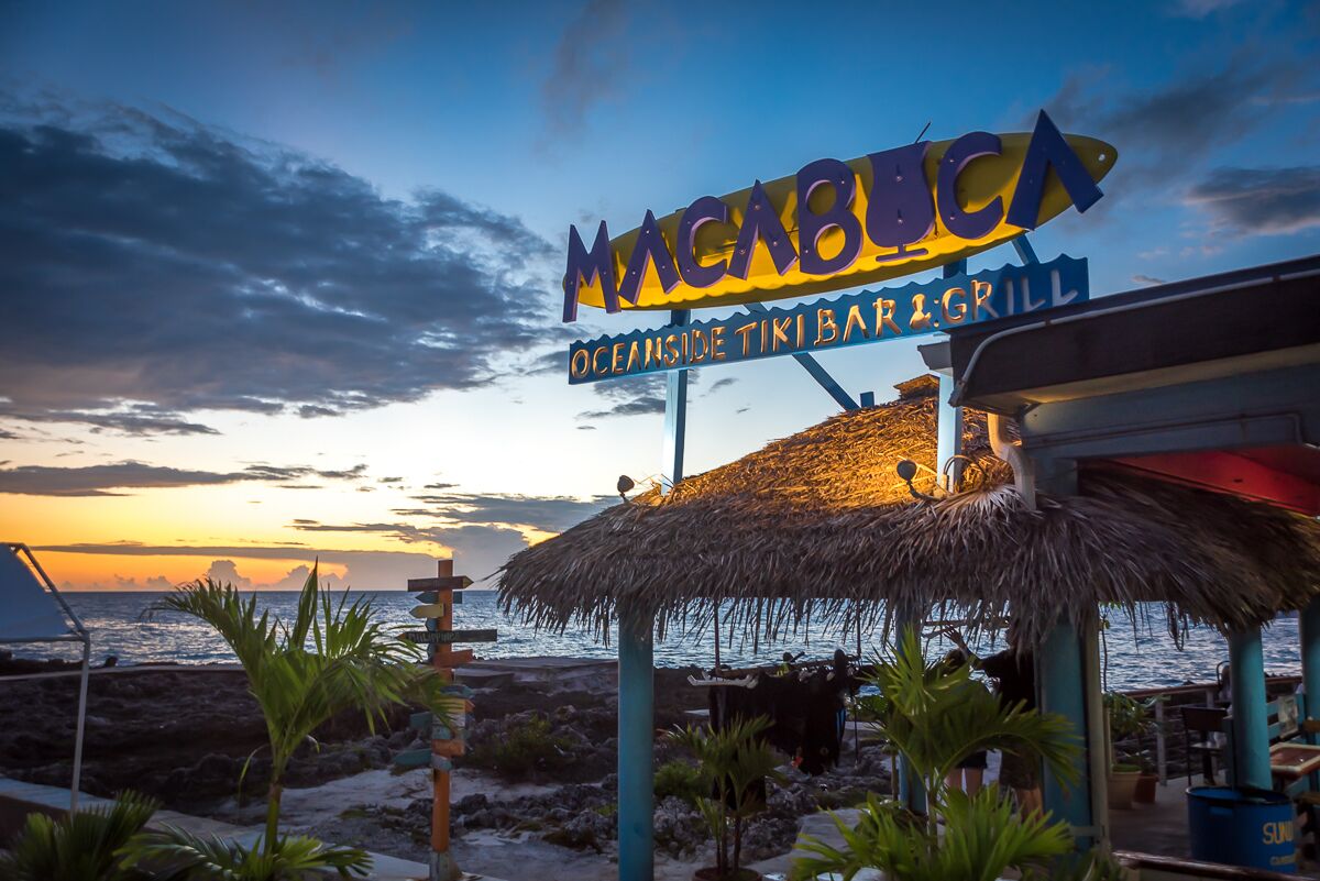 Un restaurante frente al mar llamado 'Macabucca', con techo de paja y vistas al océano al atardecer.