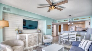 Sala de estar con sofá, sillón y televisor montado en la pared. Se observa un ventilador de techo y una zona de cocina al fondo.