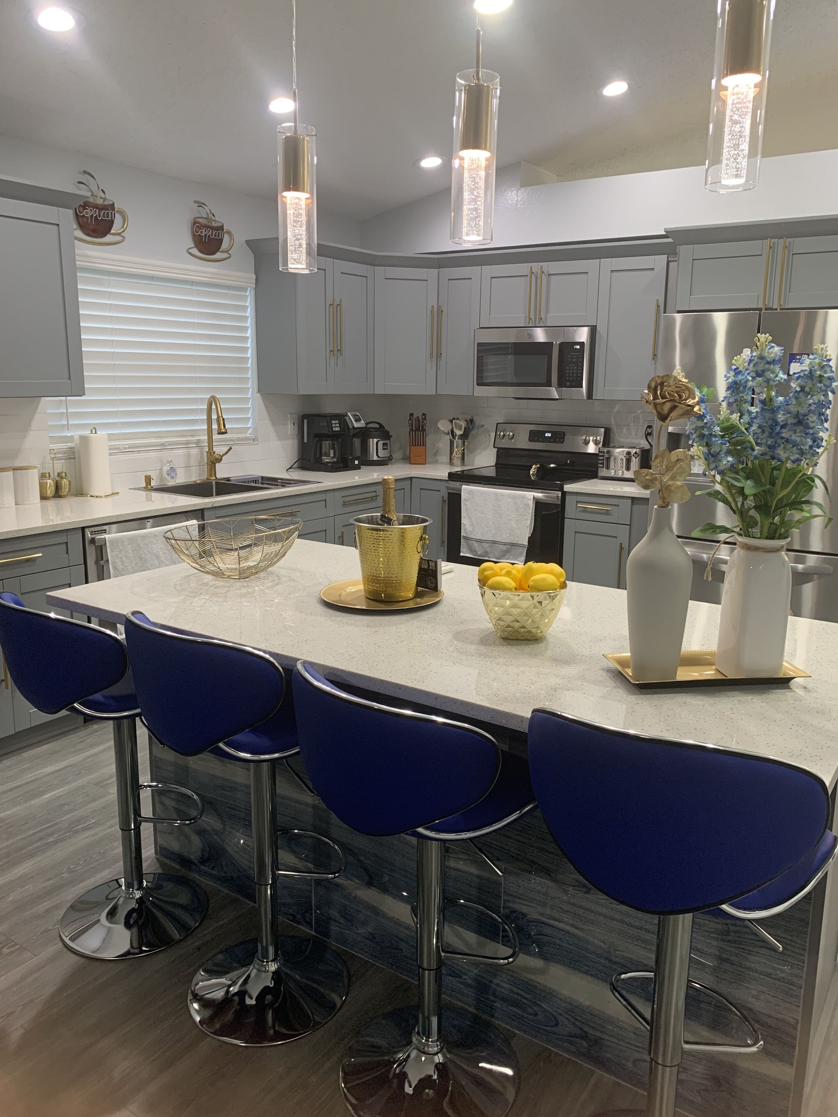 Uma cozinha moderna com armários cinza, uma ilha central com banquetas, eletrodomésticos de aço inoxidável e objetos decorativos como um vaso com flores e uma xícara de café.