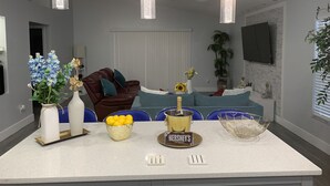 Una cocina moderna con una isla central, iluminación colgante, una zona de estar con sofá y sillas, y una mesa de comedor con un cuenco de limones y una barra de chocolate Hershey's.