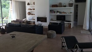 Een moderne woonkamer met een bank, voetenbankje en een eettafel. Er staan rekken met decoratieve objecten en een flatscreen televisie.
