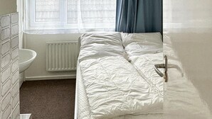 A small, narrow room with a single bed, a radiator, a window with curtains, and a sink.