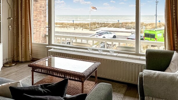 A room with a large window overlooking a beach and a road. There is a wicker table and two chairs in the room.