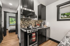 Cocina compacta con estufa, lavamanos y armarios, ventana con vista a vegetación, y puerta que da a un dormitorio.