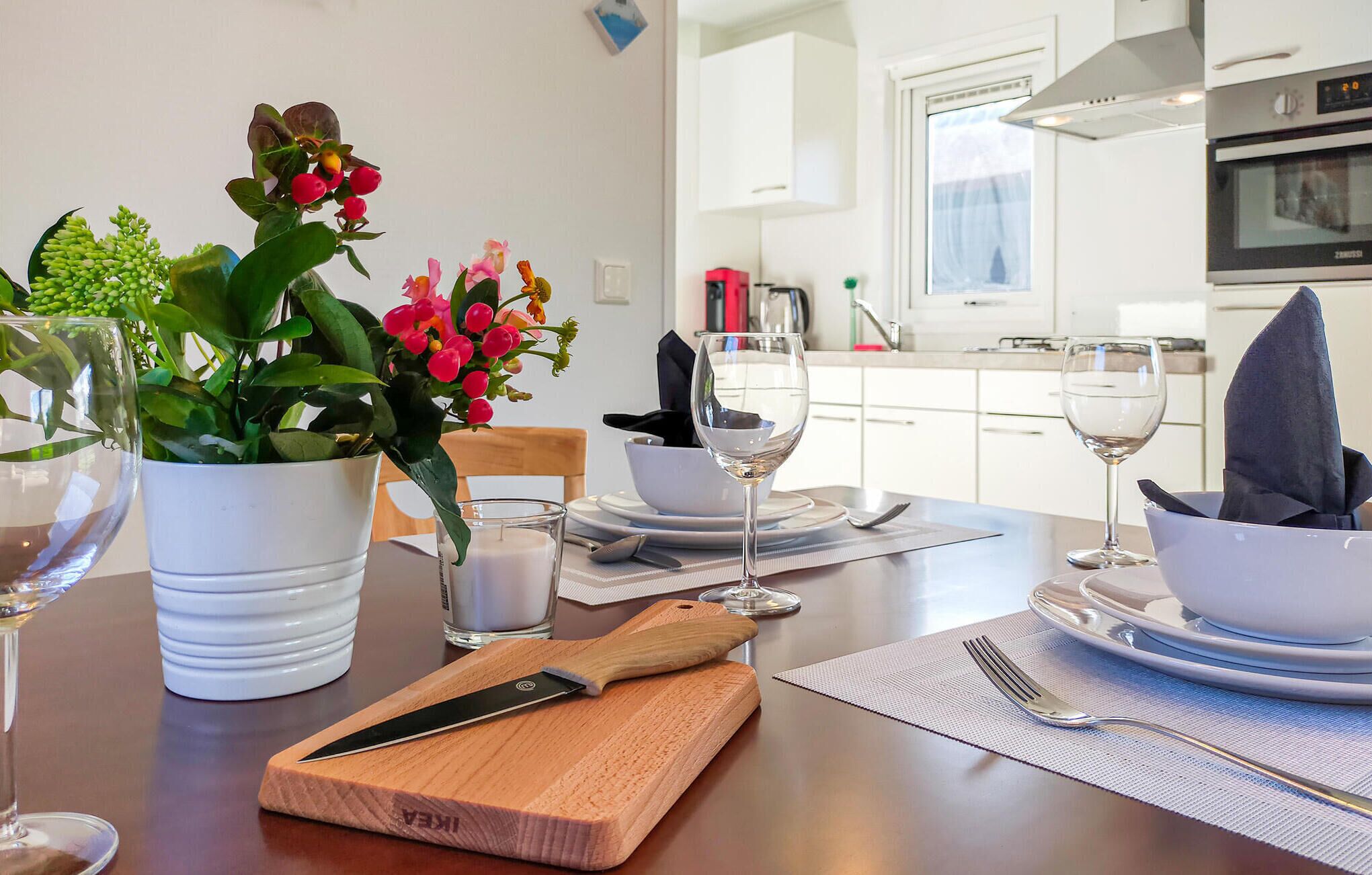 A dining table set with white plates, bowls, and cutlery, a wooden cutting board, a knife, a glass of white wine, and a potted plant with red flowers.
