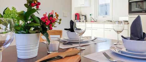 A dining table set with white plates, bowls, and cutlery, a wooden cutting board, a knife, a glass of white wine, and a potted plant with red flowers.