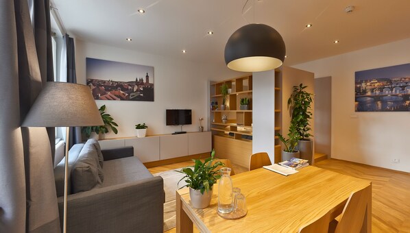 Ein modernes Wohnzimmer mit einer Couch, einem Esstisch und einem Fernseher. An den Wänden befinden sich Pflanzen, Bücherregale und Kunstwerke.
