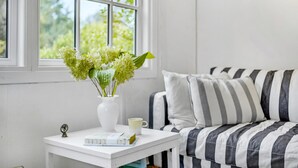 Ein ordentlich eingerichteter Raum mit einem weißen Nachttisch, einer gestreiften Couch, einem Fenster mit Grünpflanzen draußen und einer Vase mit Blumen auf dem Tisch.
