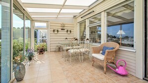 Un patio iluminado por el sol con una mesa y sillas, una silla de mimbre con cojín, un regador rosa y un jarrón grande con flores.