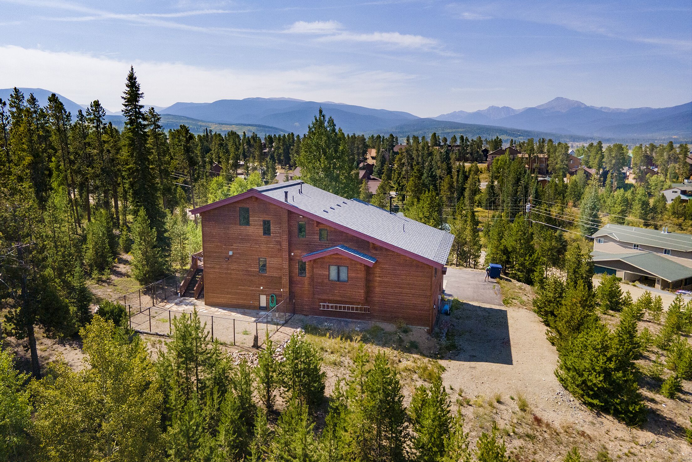 A two-story wooden cabin with a sloped roof, surrounded by dense forest and mountains in the background.