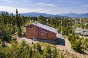 A two-story wooden cabin with a sloped roof, surrounded by dense forest and mountains in the background. - Spacious 6BR Home, Large Hot Tub, Walk to Shuttle, Attached Heated 4-Car Garage! (Fraser)