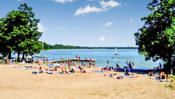 A beach with people sunbathing and swimming, a dock, and boats in the water.