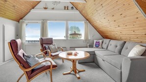 Ein gemütlicher Loftsraum mit schrägem Dach, einem großen Fenster, einer Couch, zwei Sesseln und einem runden Holztisch.