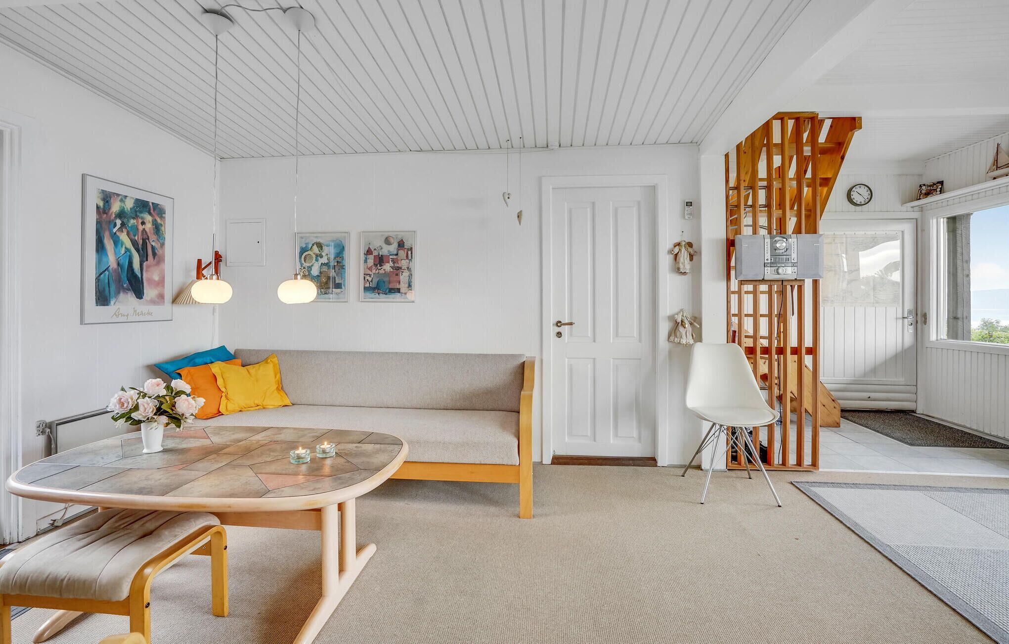 Un salón acogedor con una mesa redonda de madera, un sofá con cojines coloridos y una silla blanca. Se observan obras de arte enmarcadas en la pared y una escalera de madera al fondo.