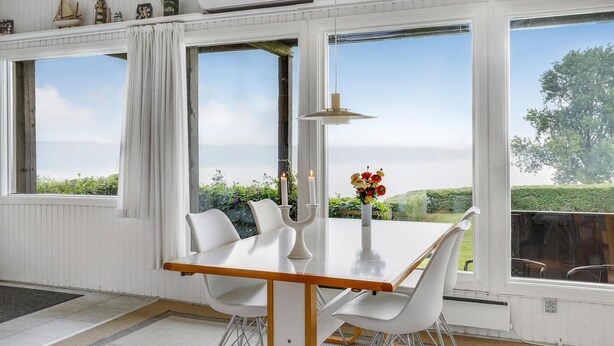 Ein Essbereich mit Tisch und Stühlen, großen Fenstern mit Blick auf Grünflächen und ein Gewässer, sowie eine Decke mit Dekorationselementen.